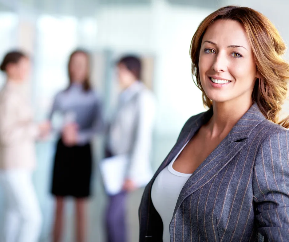 Smiling professional woman in a pinstripe blazer, with colleagues talking behind her
